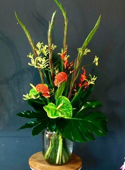 Tropical flowers to include Anthurium, Celosia, kangaroo paws, monstrea foliage in dark glasses vase